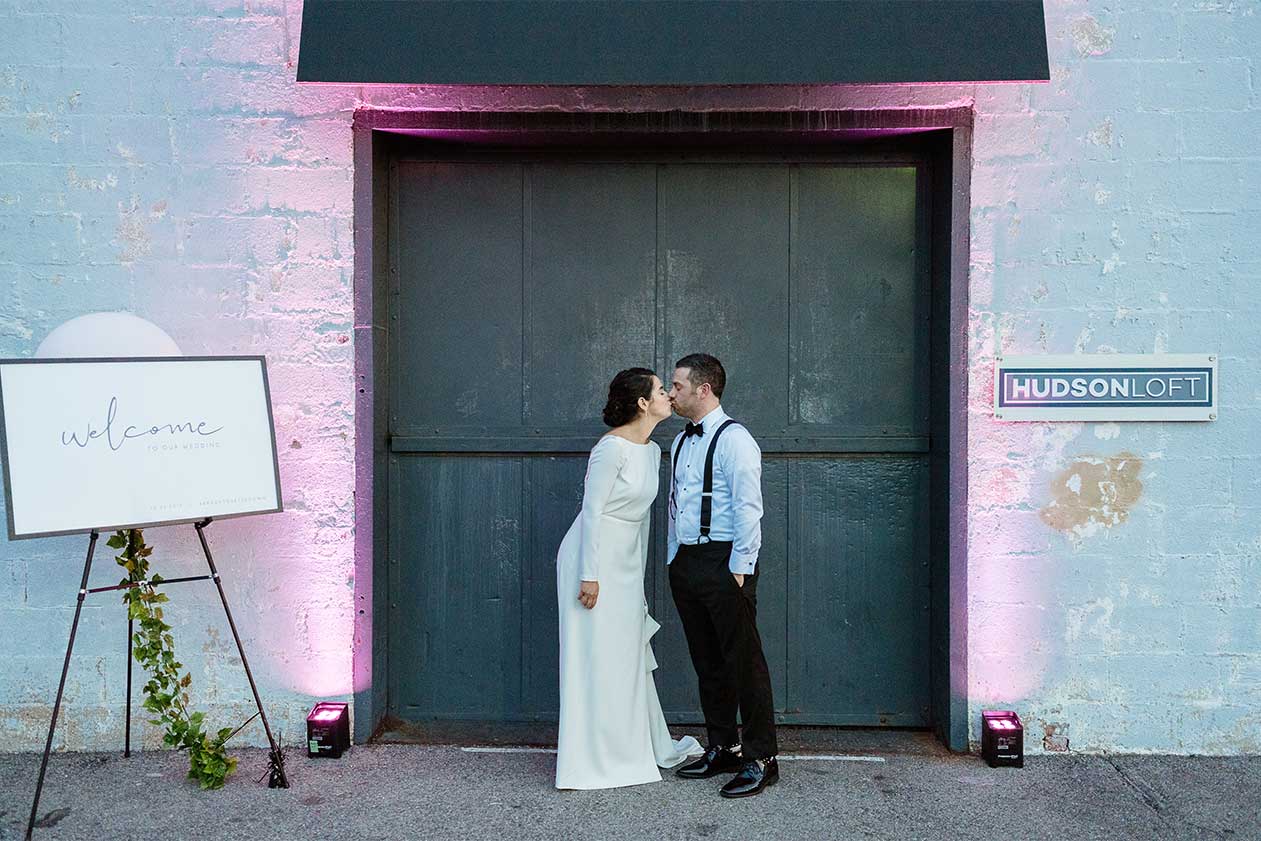 Wedding couple kissing in front of Hudson Loft’s freight elevator, next to their welcome signage with ambient up lighting